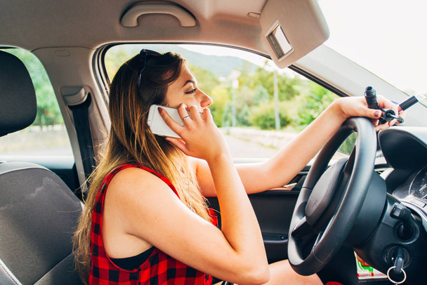 Dikkati dağılmış genç bir kadının portresi akıllı telefonuyla konuşuyor ve dikkatsizce araba kullanırken rimel sürüyor. - Fotoğraf, Görsel