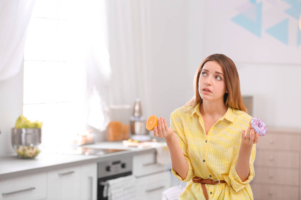Thoughtful young woman choosing between orange and donut in kitchen, space for text. Healthy diet - Photo, image