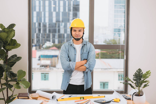 sonriente asiático arquitecto con brazos cruzados de pie en el lugar de trabajo con planos
 - Foto, Imagen