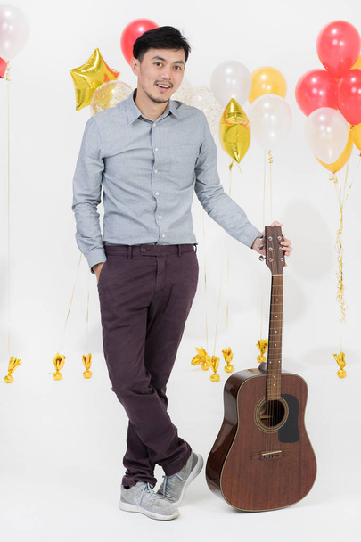 Full-length studio portrait of Asian young man, black hair, in gray shirt, smiling,look straight to camera,holding acoustic guitar on party scene with bunch of balloons on isolated white background - Photo, Image