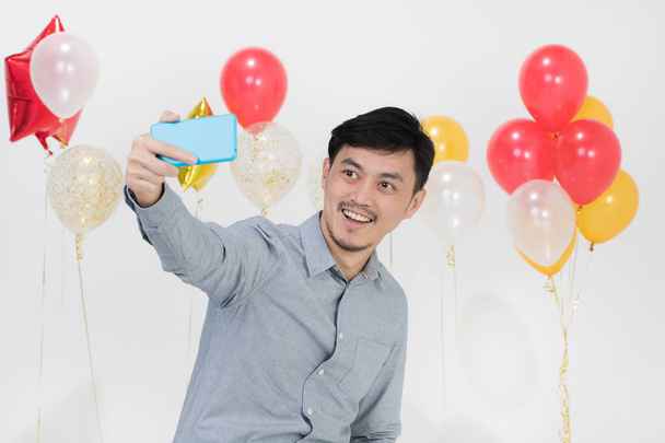 Portrait de beau jeune homme asiatique, cheveux noirs, en chemise grise, souriant joyeusement, prenant selfie avec smartphone sur scène de fête avec des grappes de ballons sur fond blanc isolé
 - Photo, image