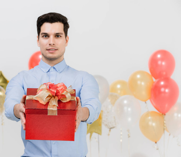 Festive gift box in focus, holded by young caucasian man  at work party, studio shot white background and festive balloons. - Photo, image