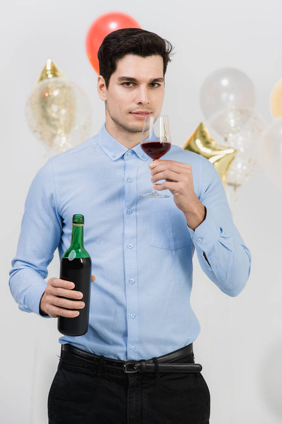 Homem caucasiano jovem atraente, tesing vinho casado de vidro sentindo-se feliz na festa de trabalho, fundo branco com balões coloridos festivos
. - Foto, Imagem