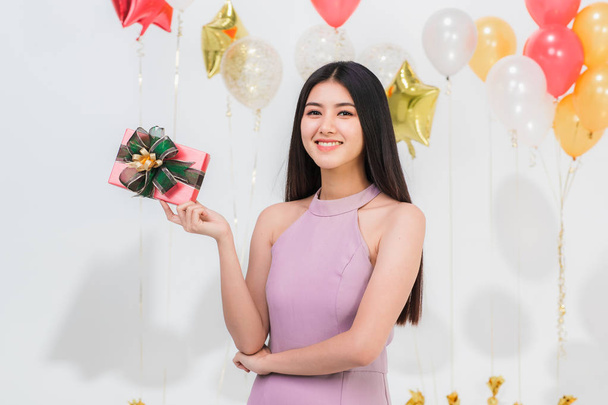 Όμορφη νεαρή γυναίκα της Ασίας μεγάλο φιλικό χαμόγελο posig με το κιβώτιο δώρων ευτυχισμένη διασκέδαση κόμμα, κατακόρυφος βολή λευκό φόντο με πολύχρωμα μπαλόνια. - Φωτογραφία, εικόνα