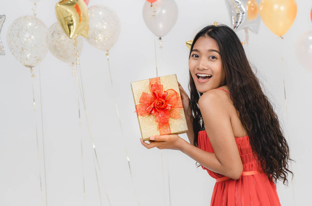 Приваблива молода азіатка носить червону сукню, схвильовану своїм подарунком, щаслива на веселій вечірці, портретний знімок білого фону зі святковою повітряною кулею
. - Фото, зображення