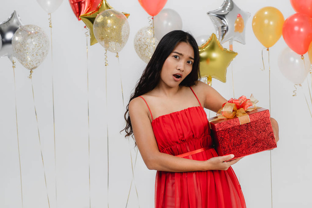 attraktive junge asiatische Frau trägt rotes Kleid begeistert von ihrem Geschenk, glücklich auf einer lustigen Party, Portraitaufnahme weißer Hintergrund mit festlichem Luftballon. - Foto, Bild