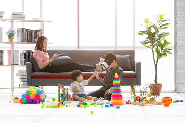 Asian family, happy time at home, young beautiful mother, relaxing, using tablet on couch, young boy wear white imitation games goggles headset playfully touch hands with little adorable toddler - Fotoğraf, Görsel