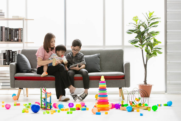 Asian family, happy, leisure time at home, young beautiful mother showing book to cute, little toddler drinking from milk bottle, young son wearing eyes glasses in her arms, in modern living room - Photo, Image