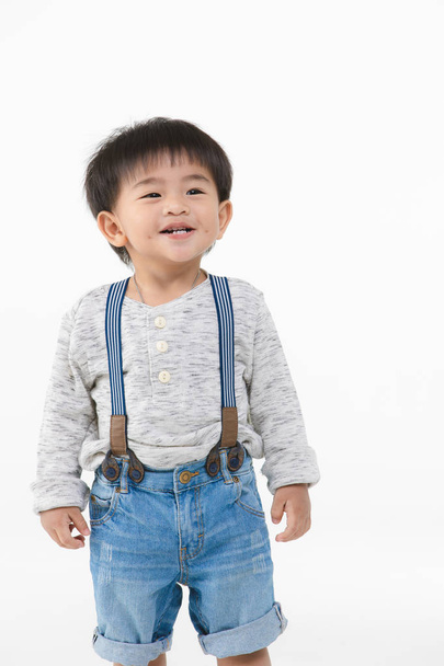 Полудлинный студийный портрет милого, очаровательного азиатского мальчика в джинсовом комбинезоне, футболка с длинным рукавом, счастливо улыбающегося, на изолированном белом фоне
 - Фото, изображение