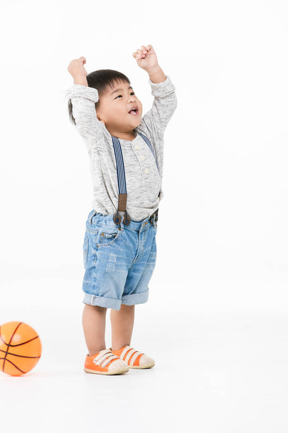 Studio portrait of cute, adorable, Asian toddler boy wearing denim overalls, long sleeve T-shirt, orange shoes, standing, smiling, hands over his head, orange ball behind, on isolated white background - Foto, Imagen