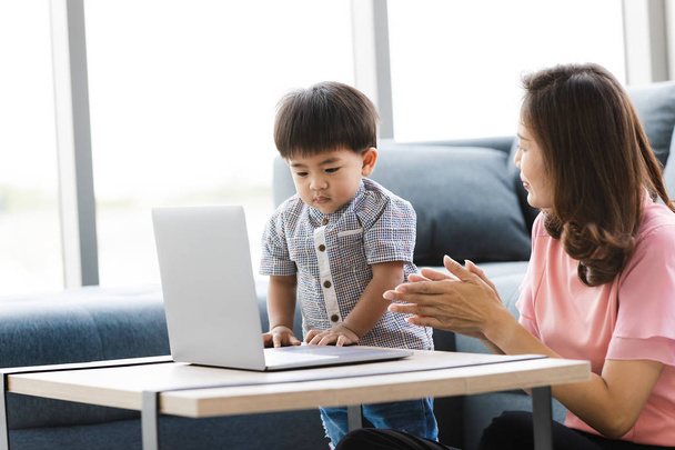 Азиатская семья, счастливое время дома, молодая красивая мать, сидящая на полу, симпатичный, обожаемый малыш, на своих лапах, ласково указывая ему что-то на экране ноутбука, на фоне дивана
 - Фото, изображение
