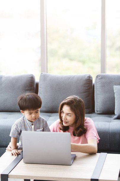 Азіатський сім'ї, щасливий час вдома, молода мати красиві, сидячи на підлозі, милий, чарівні малюк на її кіл, як Ласкаво вказуючи щось на екрана ноутбука для нього, з фоном диван - Фото, зображення