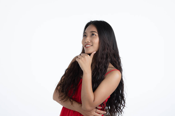 Студийный портрет молодой красивой азиатки, длинные кудрявые черные волосы, скрещенные руки, рука на подбородке, улыбка и взгляд вверх в мыслящей позе, на изолированном белом фоне
 - Фото, изображение