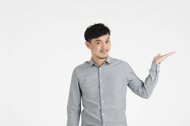 Полудлинный студийный портрет молодого черноволосого азиата в серой рубашке, протягивает левую руку, раскрывает ладонь, как будто держит что-то воображаемое на изолированном белом фоне
 - Фото, изображение