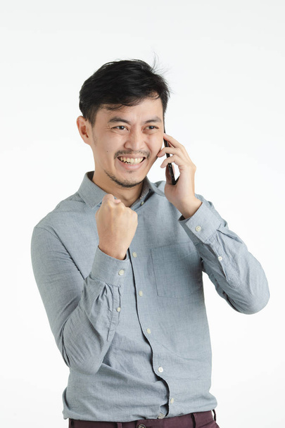 Portrait studio mi-long de jeune homme asiatique en chemise grise, les poings serrés, expression enthousiaste sur le succès, sourire, parler sur un téléphone portable, regarder loin de la caméra sur fond blanc isolé
 - Photo, image