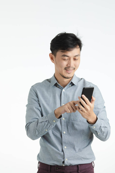 Portrait studio mi-long de cheveux noirs, jeune homme asiatique en chemise grise, au visage souriant, tenant et utilisant un smartphone, sur fond blanc isolé
 - Photo, image