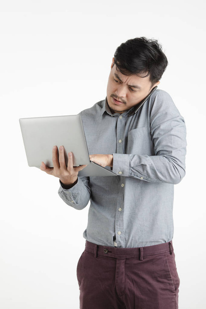 halblanges Studioporträt schwarzer Haare, junger asiatischer Mann im grauen Hemd, hält und tippt auf digitalem Notizbuch, spricht auf Smartphone, auf isoliertem weißem Hintergrund - Foto, Bild
