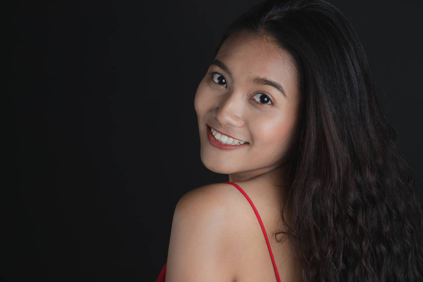Half-length studio portrait of young beautiful,long,curly black hair Asian woman in red bare shoulder dress,turning back,tilted head,looking away from camera, on dark background - Photo, Image