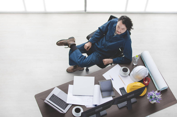 Top view of Asian beard male foreman, in coverall, wear safety shoes, relax, sit on chair, coffee cup on table, with stationery, safety helmets, laptop, tablet, smartphone, office background - Photo, Image