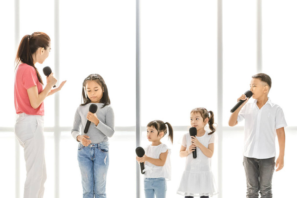 asiatische Lehrer bringen asiatischen Kindern bei, ein Lied zu singen, Kinder achten darauf, ihr zuzuhören, sie stehen vor einem großen weißen Fenster. Konzept für Musikschule oder Gesangsunterricht. - Foto, Bild