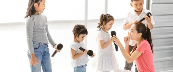 Asian teacher teach Asian kids to sing a song, kids pay attention to listen to her, they stand in front of big white window. Concept for music school or singing classroom training. - 写真・画像