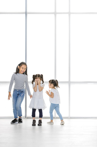 Asian kids point at Asian girl, the stand beside big white window. - Photo, Image