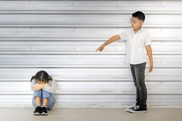 Asian boy point at sitting Asian girl, white wall behind them. - 写真・画像