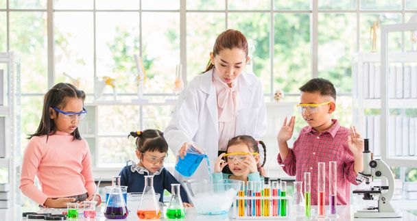 Naturwissenschaftler unterrichten asiatische Schüler über Chemikalien, Lehrer gießen Chemikalien in Glasschale, buntes Reagenzglas und Mikroskop auf Tisch im Laborraum, Konzept für das Lernen im Laborraum. - Foto, Bild