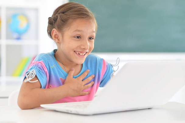 Емоційна маленька дівчинка використовує ноутбук, сидячи за столом у класі
 - Фото, зображення