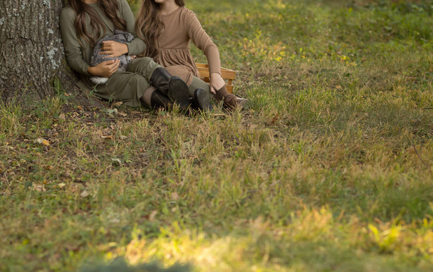 dos chicas se sientan debajo de un árbol en la hierba y acarician un conejo gris. tonos pastel de beige y verde. otoño o primavera
 - Foto, Imagen