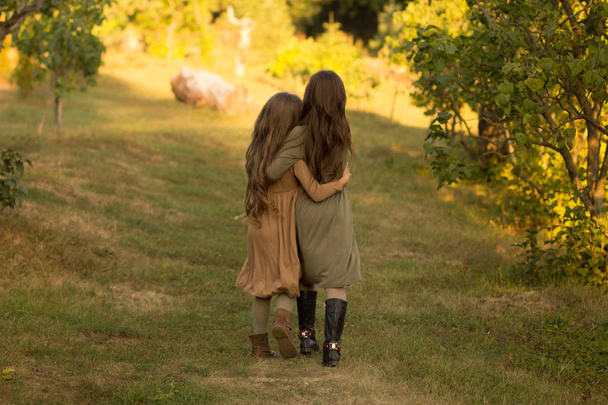 two girls hug on the street in the forest. friendship. in green and brown dress. a park. autum.back view - Foto, afbeelding