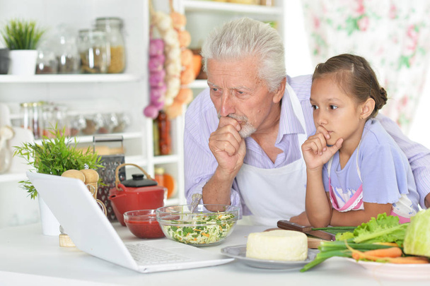 Senior man with granddaughter looking at laptop while preparing dinner in kitchen - Foto, afbeelding