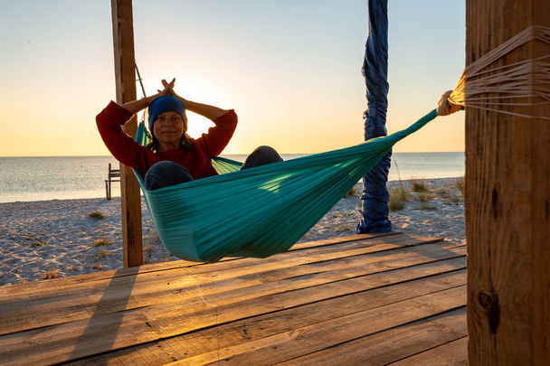 Smiling woman relaxes in a hammock on abandoned bungalow veranda, admiring the turquoise sea during sunset and meditates. Awesome travel to the seaside. Backlight. - Zdjęcie, obraz