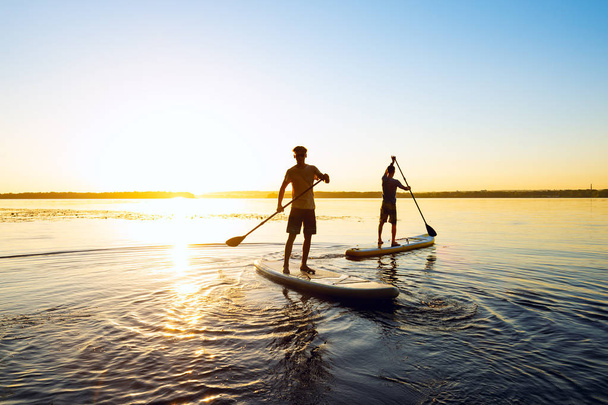 Men, friends are paddling on a SUP boards on a large river during sunrise. Stand up paddle boarding - awesome active recreation in nature. Backlight.  - Fotó, kép