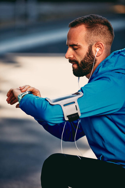 Bearded man kneeling on the street and checking heart beat on smart watch. Headphones in ears. Healthy lifestyle concept. - Foto, afbeelding