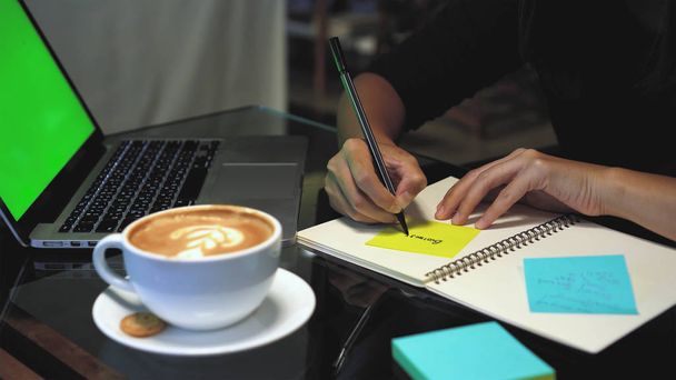 Closeup hands of Asian businesswoman working and writing on paper Notes over the notebook with green screen laptop and coffee cup in coffee cafe, Lifestyle and business concept - Photo, Image