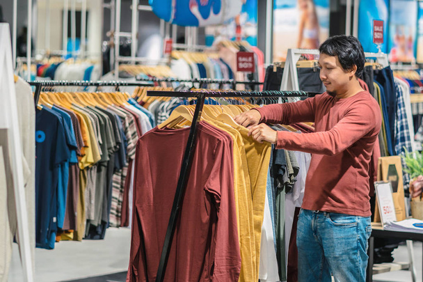 Inteligentny człowiek Azjatka z brodą, wybierając ubrania w sklepie odzieżowym w centrum handlowe, patrząc kolorowe t-sshirts, mody i koncepcja konsumpcjonizm. - Zdjęcie, obraz