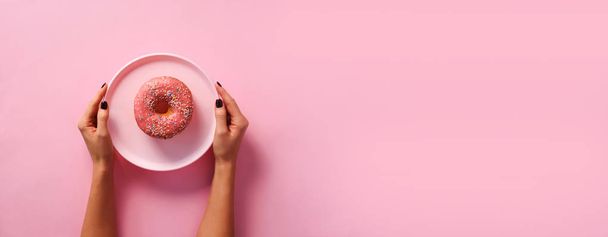 Female hands holding donut on plate over pink background. Top view, flat lay. Sweet, dessert, diet concept. Banner with copy space. Weight lost after holidays. - Foto, imagen