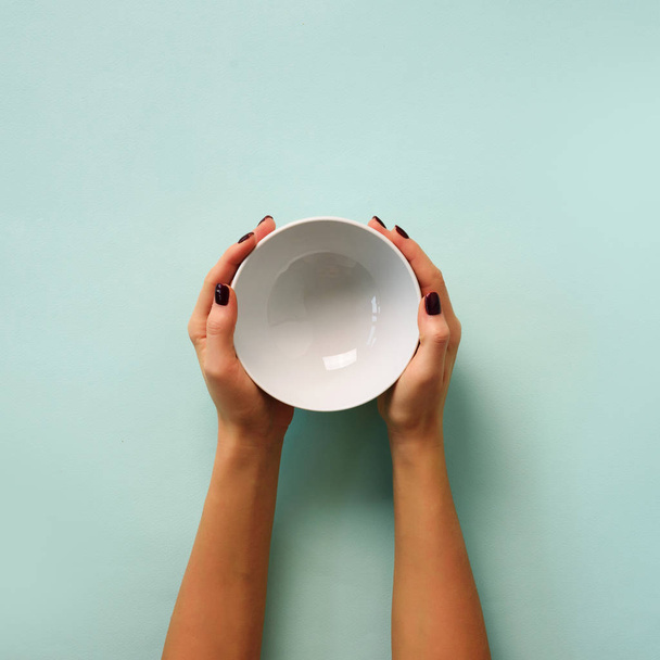 Female hand holding white empty bowl on blue background with copy space. Healthy eating, dieting concept. Banner. Square crop. - Φωτογραφία, εικόνα