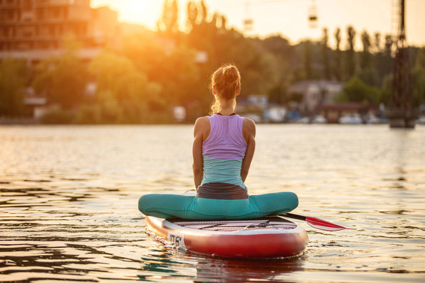 Young woman doing yoga on sup board with paddle. Yoga pose, side view - concept of harmony with the nature. - Photo, Image