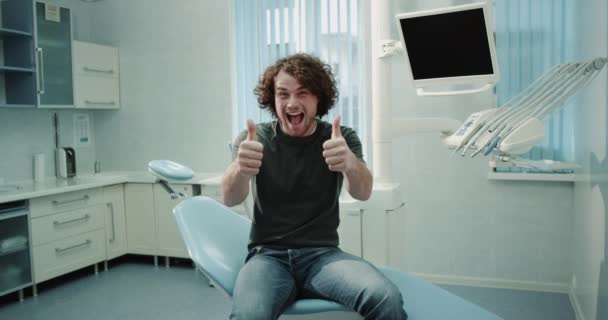 Young man in a dental clinic smiling large in front of the camera and showing a big like after his hygiene checking , sitting on a blue dentist chair. - Materiaali, video