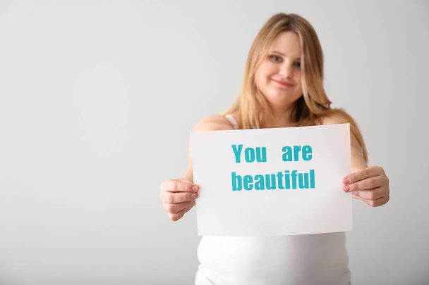 Beautiful plus size girl holding paper sheet with text YOU ARE BEAUTIFUL on light background. Concept of body positivity - Foto, Imagem
