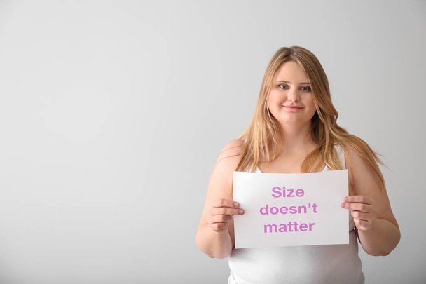 Beautiful plus size girl holding paper sheet with text SIZE DOESN'T MATTER on light background. Concept of body positivity - Foto, immagini