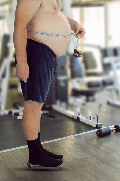 Fat gut middle age man in trunks and black socks stands on the scale and measures his waist with a measuring tape in order to claim that he has not lost weight and waist after the exercising in the gym and receive his money back. - Фото, изображение