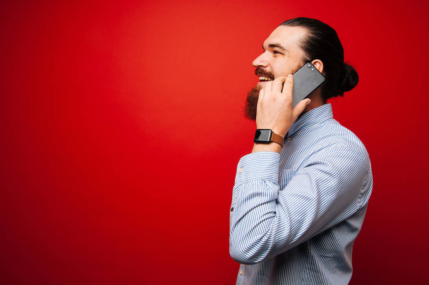 glücklicher junger bärtiger Mann in lässigem Lächeln und Gespräch auf Smartphone über rotem Hintergrund - Foto, Bild