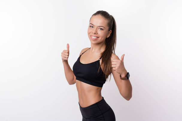 Charming and smiling young fitness woman showing thumbs up over white background - Foto, immagini