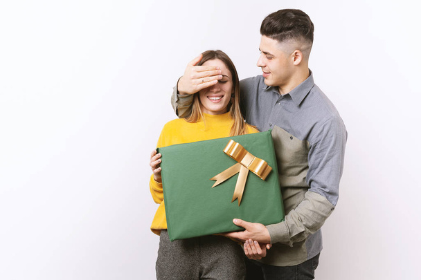 白い背景の上の幸せな若いカップル。男はプレゼントを与える彼女 ang に目を覆っています。クリスマスやバレンタインデーのコンセプト. - 写真・画像