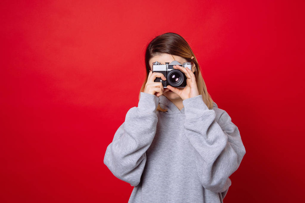 Молодая модная женщина стоит на красном фоне и фотографируется со старой винтажной ретро-камерой
 - Фото, изображение