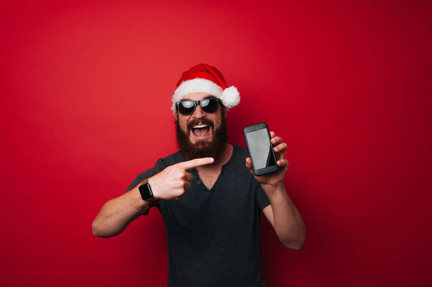 Cheerful happy bearded man wearing santa claus hat pointing at smartphone over red background - Photo, Image