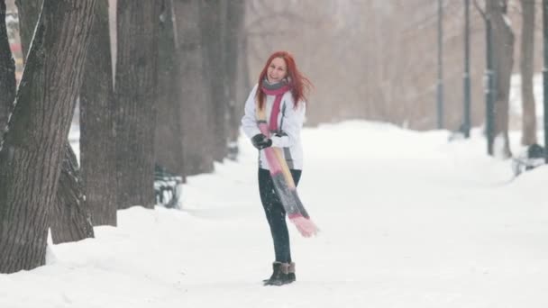 Talvipuistossa. Iloinen nainen, jolla on kirkkaan punaiset hiukset, seisoo jalkakäytävällä, tekee lumipallon ja heittää sen eteenpäin
 - Materiaali, video